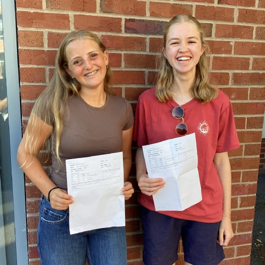 two sixth form students smiling with their A level results in their hands